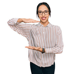 Wall Mural - Young hispanic girl wearing casual clothes and glasses gesturing with hands showing big and large size sign, measure symbol. smiling looking at the camera. measuring concept.