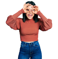 Poster - Young brunette woman with blue eyes wearing elegant sweater doing ok gesture like binoculars sticking tongue out, eyes looking through fingers. crazy expression.