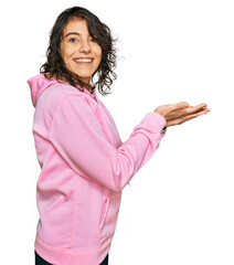 Wall Mural - Young hispanic woman wearing casual sweatshirt pointing aside with hands open palms showing copy space, presenting advertisement smiling excited happy