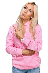 Wall Mural - Young blonde woman wearing casual sweatshirt pointing with hand finger to the side showing advertisement, serious and calm face