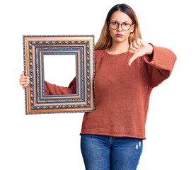 Poster - Beautiful young woman holding empty frame with angry face, negative sign showing dislike with thumbs down, rejection concept