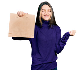 Wall Mural - Young brunette girl with long hair holding take away paper bag screaming proud, celebrating victory and success very excited with raised arm