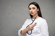 Beautiful female doctor holding stethoscope hearing heart beating indoors. Portrait of serious woman wearing uniform using stethoscope for herself diagnostic, against grey wall. Medicine concept.