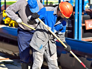 Two workers paint metal pipe black with paint rollers. Pipe treatment for corrosion protection. Metal paint. Two handymen paint gasification pipe on summer day.