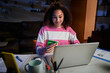 © CarlosBarquero - Smiling latina woman using laptop shopping in online store from home at night. Young afro female sitting in lounge with computer and credit card in hand. Internet sales, adding to discounts at Gen z