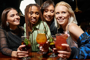  joyful and elegant female friends holding glasses with delicious cocktails in bar, friendship