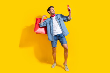 Full body photo of cool man dressed denim shirt shorts holding new outfit in bags look at smartphone isolated on yellow color background.