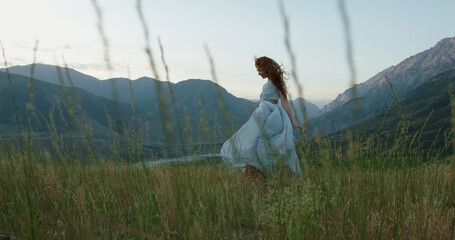 Wall Mural - Girl in light dress with red hair is walking on top of a spring mountain with scenic view. Woman in search for inspiration - freedom concept 