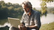 An elderly gray-bearded man with a laptop and a smartphone works in the summer on the street by the river, the concept of an open-air office