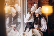 Two Girls Sisters With Long Hair In White Hats And White Cardigans And Jeans Sitting At The Window And Looking Outside. Christmas Tree . Winter Time