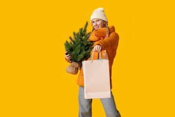 Poster - Young woman in warm winter clothes with shopping bag and Christmas tree on yellow background