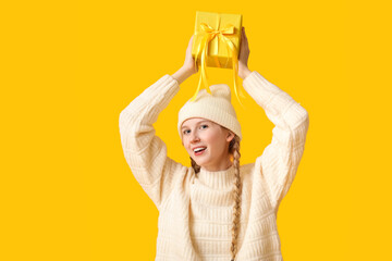 Wall Mural - Beautiful young woman in warm winter clothes with gift box on yellow background