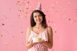 Beautiful young woman in party hat with sweet cake and confetti celebrating Birthday on pink background
