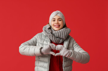 Wall Mural - Beautiful young happy woman in warm winter clothes showing heart gesture on red background