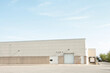 © Penguin - white doors and receiving bay doors on large one story beige building with large empty parking cement lot pavement space in front, bottom of frame, medium wide shot