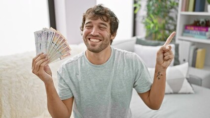 Sticker - Cheerful young man at home, holding colombian pesos, confidently pointing to the side with a joyful smile