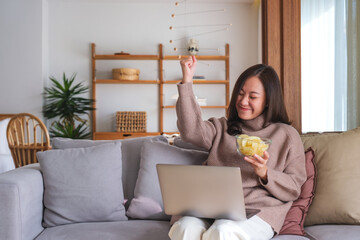 Wall Mural - Portrait image of a happy woman eating potato chips while working or watching movie on laptop at home