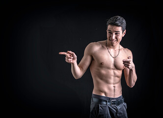 Wall Mural - Handsome shirtless young man laughing and pointing finger to a side, on black background