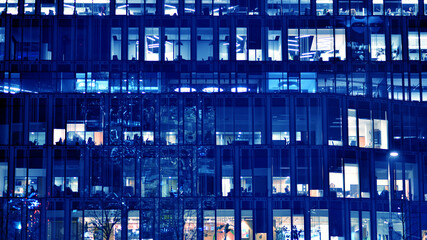 Fragment of the glass facade of a modern corporate building at night. Big glowing windows in modern office buildings at night, in rows of windows light shines. Blue graphic filter.