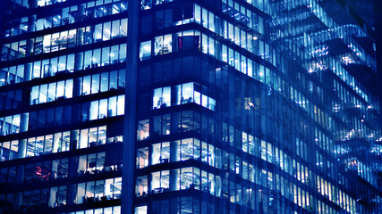 Fragment of the glass facade of a modern corporate building at night. Big glowing windows in modern office buildings at night, in rows of windows light shines. Blue graphic filter.