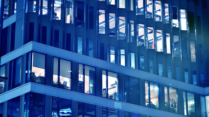 Fragment of the glass facade of a modern corporate building at night. Big glowing windows in modern office buildings at night, in rows of windows light shines. Blue graphic filter.