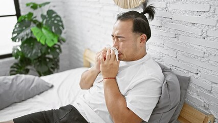 Wall Mural - Sick young chinese man, suffering from covid-19 infection, sitting on bed, sneezing in his bedroom, fighting the flu with the support of healthcare