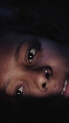 Vertical Close-up of young African American woman relaxing looking at cell phone in dark. Black girl reading or watching something on mobile lying indoors. Face illuminated by light from device screen