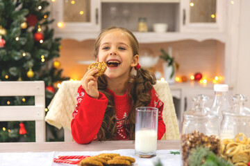 Happy joyful cute little girl in red sweater drinking milk and eating delicious cookie near Christmas tree in the kitchen for Christmas morning. Christmas time. Christmas holidays