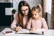 © Stavros - Mom helps daughter do homework sitting at table writing in notebook right answer. Girl asks mother to help with homework at table with laptop. Mom in cozy modern apartment helps daughter with lessons