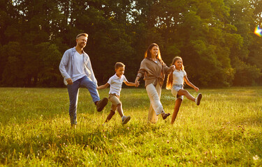 Wall Mural - Happy smiling family of four with two kids boy and girl walking in summer park enjoying beautiful nature. Young parents and their children holding hands outdoors enjoying time together.