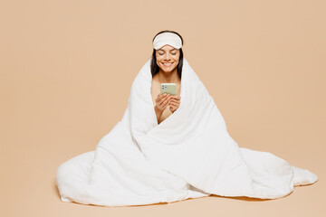 Wall Mural - Full body young happy calm Latin woman wrapped in duvet wears jam sleep eye mask rest relax at home hold use mobile cell phone isolated on plain pastel beige background. Good mood night nap concept.