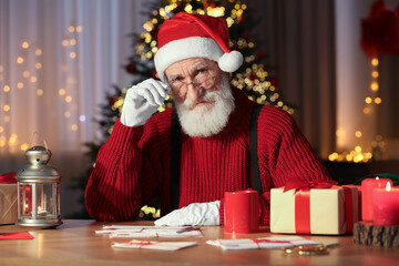 Poster - Santa Claus at his workplace. Letters, cup of beverage and gift boxes on table in room with Christmas tree