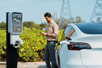 Man pay for electricity with smartphone while recharge EV car battery at charging station connected to power grid tower electrical as electrical industry for eco friendly car utilization.Expedient