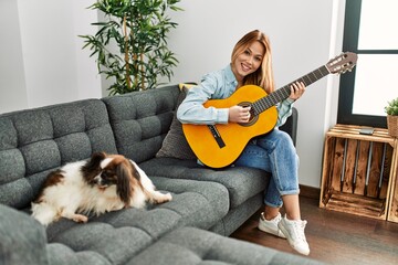 Wall Mural - Young caucasian woman playing classical guitar sitting on sofa with dog at home