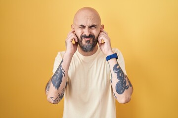 Poster - Hispanic man with tattoos standing over yellow background covering ears with fingers with annoyed expression for the noise of loud music. deaf concept.