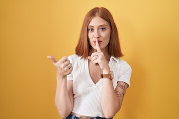 Canvas Print - Young redhead woman standing over yellow background asking to be quiet with finger on lips pointing with hand to the side. silence and secret concept.