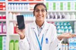 © Krakenimages.com - Young hispanic woman working at pharmacy drugstore showing smartphone screen celebrating achievement with happy smile and winner expression with raised hand