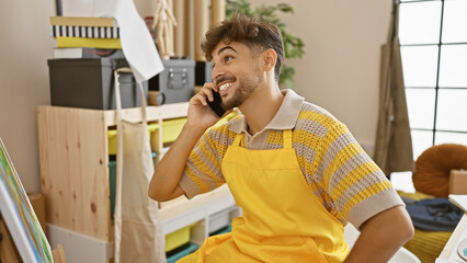 Poster - Confident arab man, a smiling art student, excitedly talking on his smartphone amidst paints and canvases in his vibrant university studio.