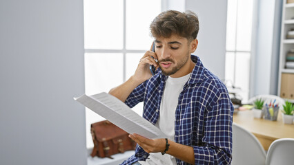 Poster - Focused young arab man, a business worker, engrossed in reading a document while having a serious conversation on his smartphone at the office