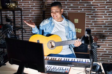 Wall Mural - Hispanic young man playing classic guitar at music studio smiling cheerful presenting and pointing with palm of hand looking at the camera.