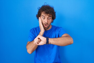 Poster - Hispanic young man standing over blue background looking at the watch time worried, afraid of getting late