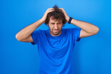 Wall Mural - Hispanic young man standing over blue background suffering from headache desperate and stressed because pain and migraine. hands on head.