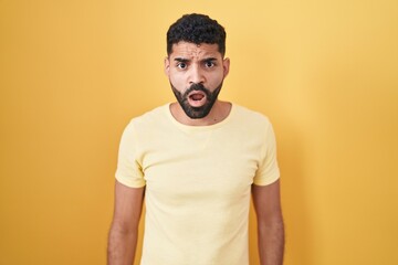 Sticker - Hispanic man with beard standing over yellow background in shock face, looking skeptical and sarcastic, surprised with open mouth