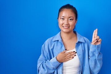 Sticker - Asian young woman standing over blue background smiling swearing with hand on chest and fingers up, making a loyalty promise oath