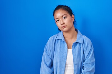 Poster - Asian young woman standing over blue background looking sleepy and tired, exhausted for fatigue and hangover, lazy eyes in the morning.