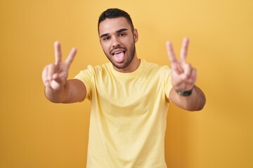 Poster - Young hispanic man standing over yellow background smiling with tongue out showing fingers of both hands doing victory sign. number two.