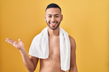 Poster - Young hispanic man standing shirtless with towel pointing aside with hands open palms showing copy space, presenting advertisement smiling excited happy