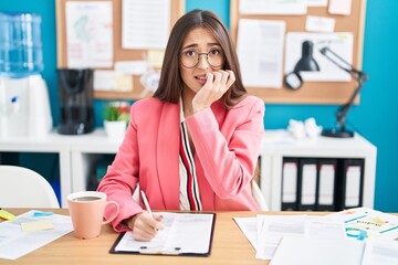 Canvas Print - Young hispanic woman working at the office wearing glasses looking stressed and nervous with hands on mouth biting nails. anxiety problem.