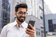 © Liubomir - Young successful businessman walking down the street holding a phone in his hands, an Indian man using an application on a smartphone smiling contentedly from outside an office building.