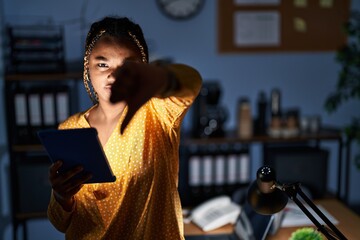 Wall Mural - African american woman with braids working at the office at night with tablet looking unhappy and angry showing rejection and negative with thumbs down gesture. bad expression.